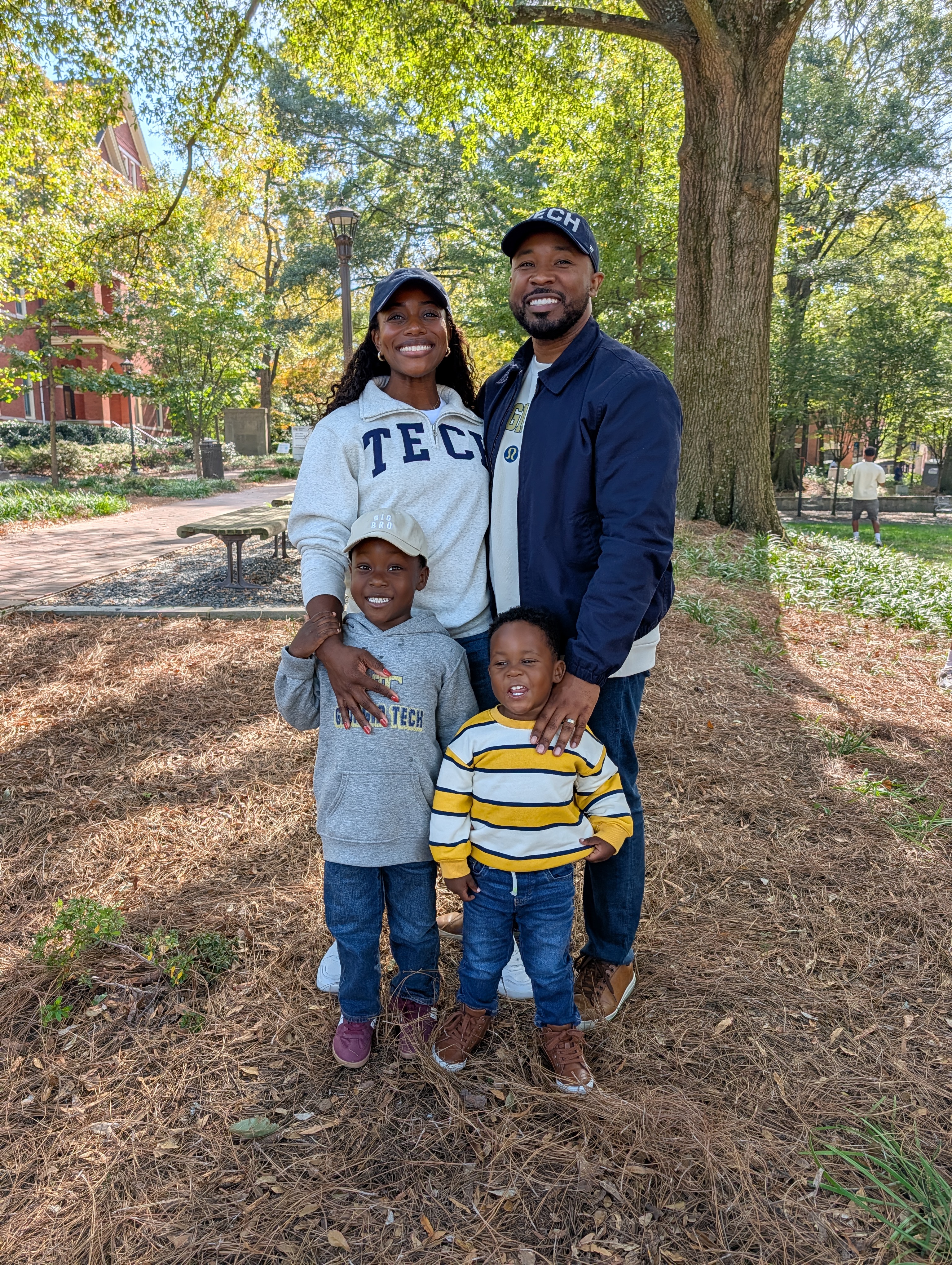 A family with two young boys stands in front of a tree.