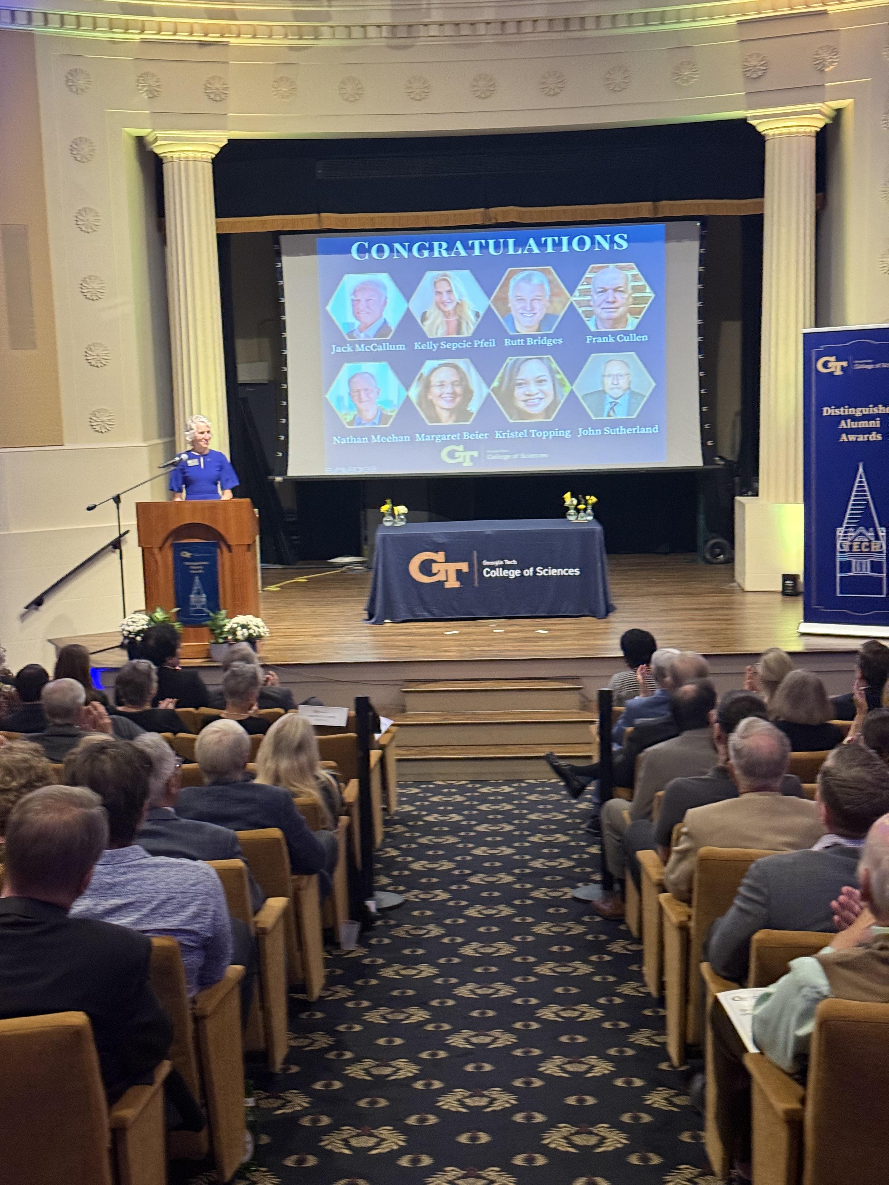 a woman at a podium in front of a screen highlighting all of the Distinguished Alumni Award winners.