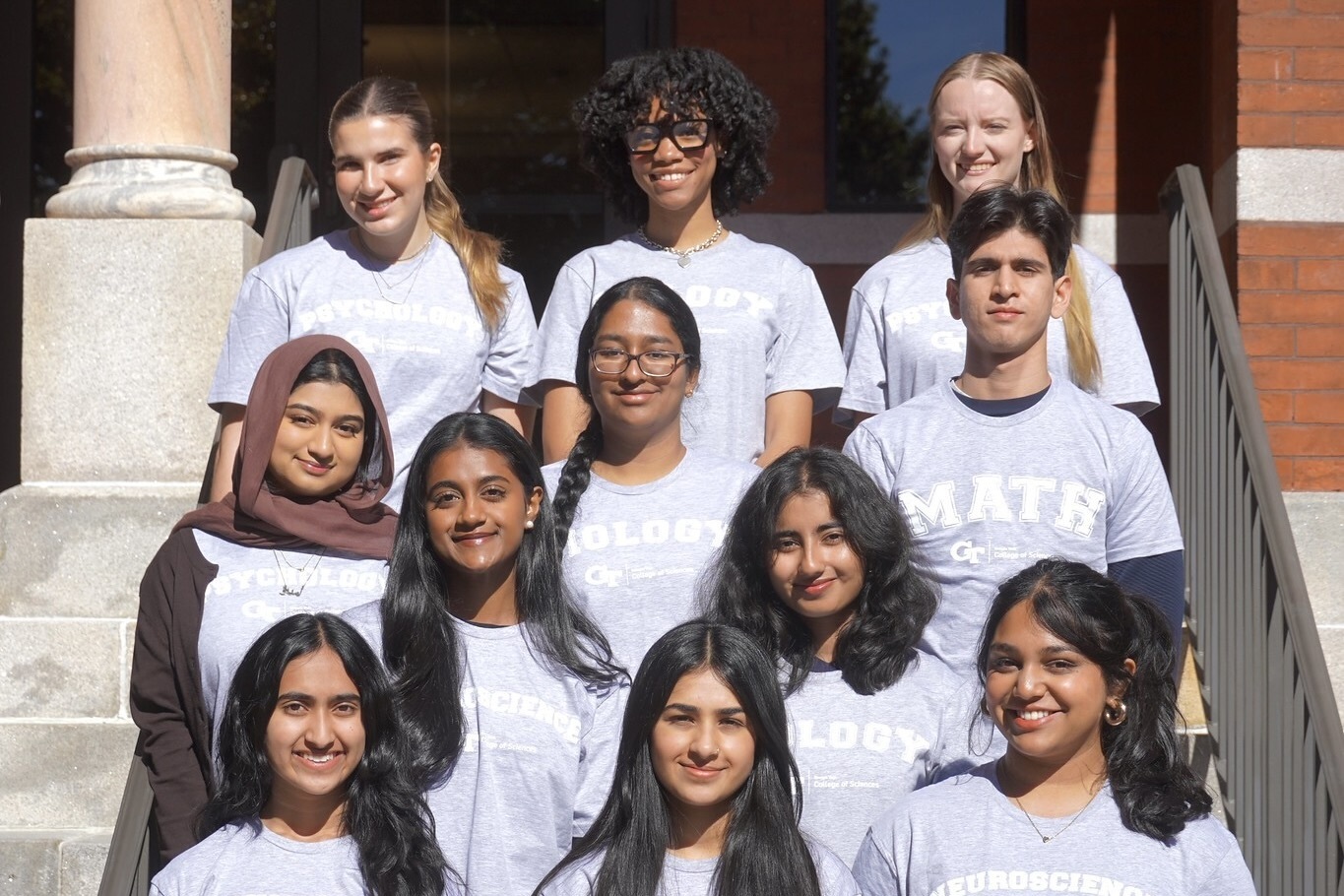 Eleven students sitting in rows in front of a brick building.